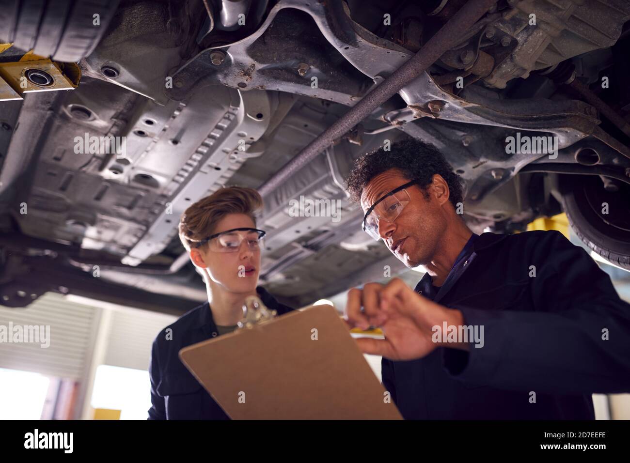 Male Tutor With Student Looking Underneath Car On Hydraulic Ramp On