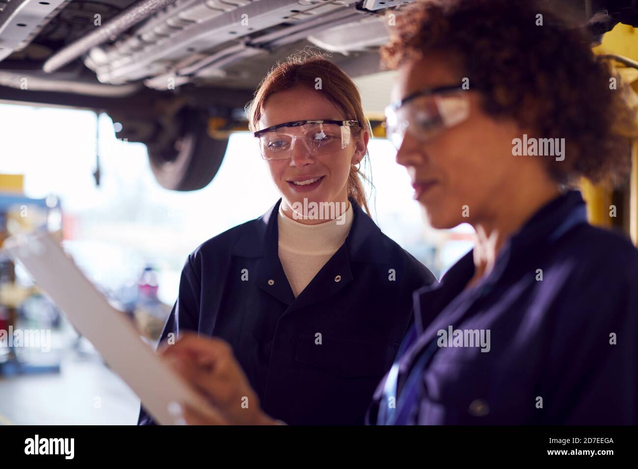 Female Tutor With Student Looking Underneath Car On Hydraulic Ramp On