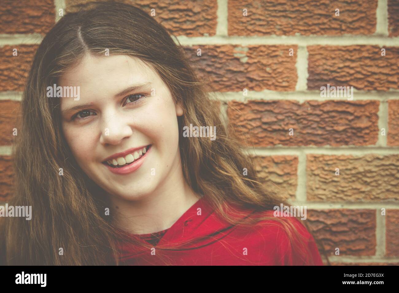 Beautiful natural portrait of teenage girl with long brown hair looking at camera with smile showing teeth against brick wall Stock Photo