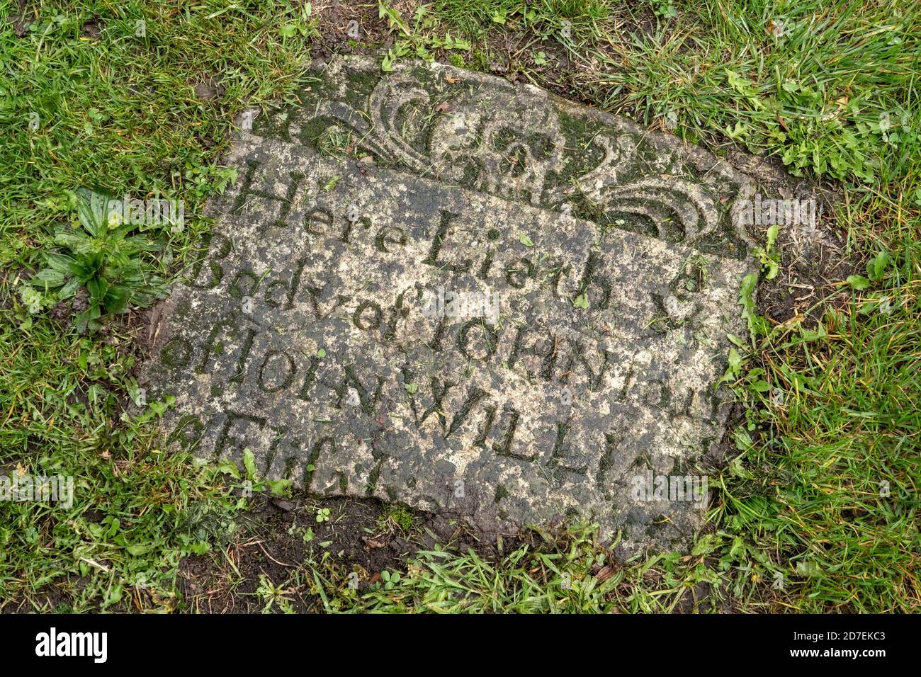 Section of graveyard headstone partly buried and overgrown by grass with skull motif visible Stock Photo