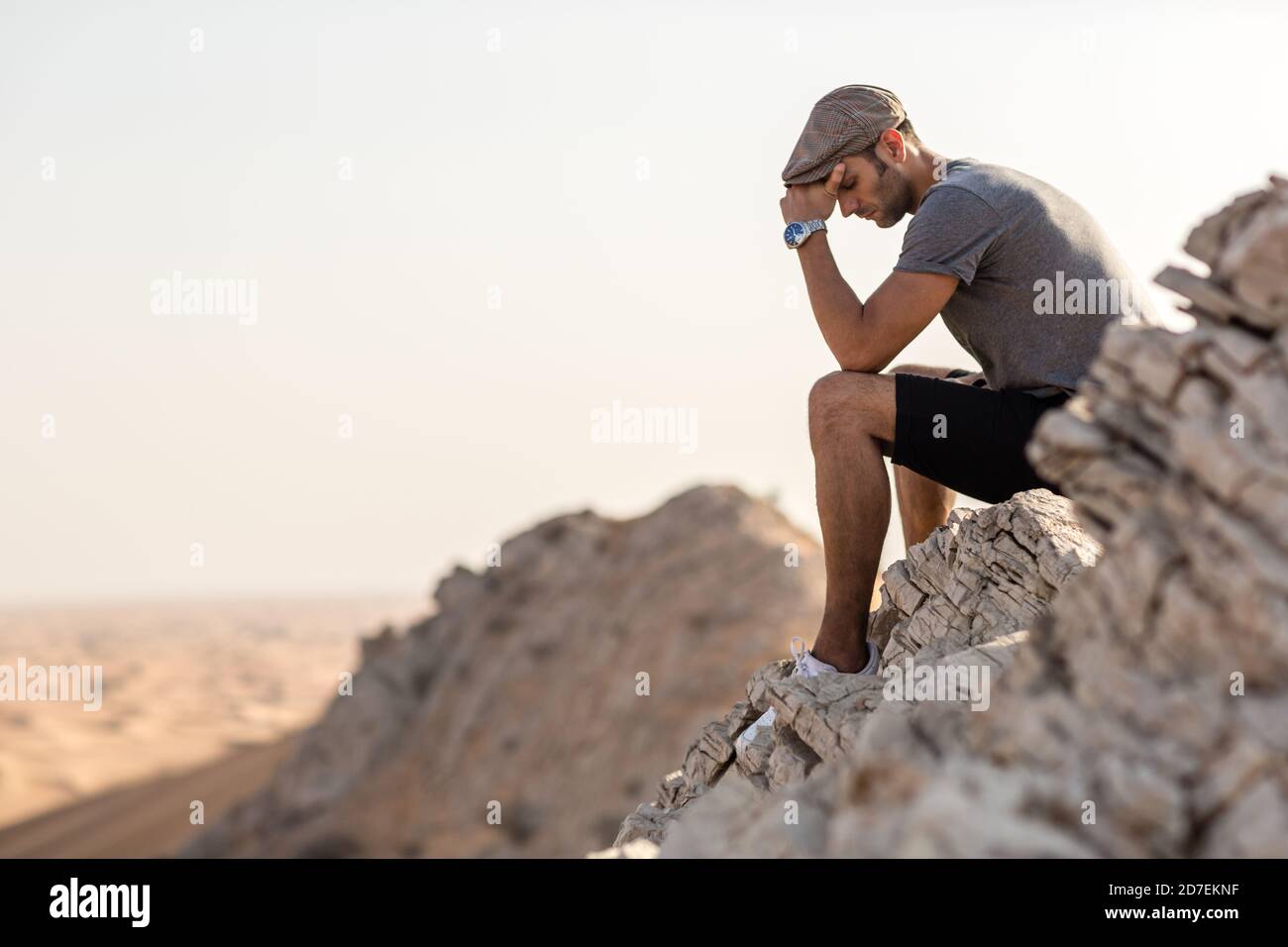 Depression and loneliness. Sad young man sitting alone on top of a mountain cliff overlooking the desert. Stock Photo