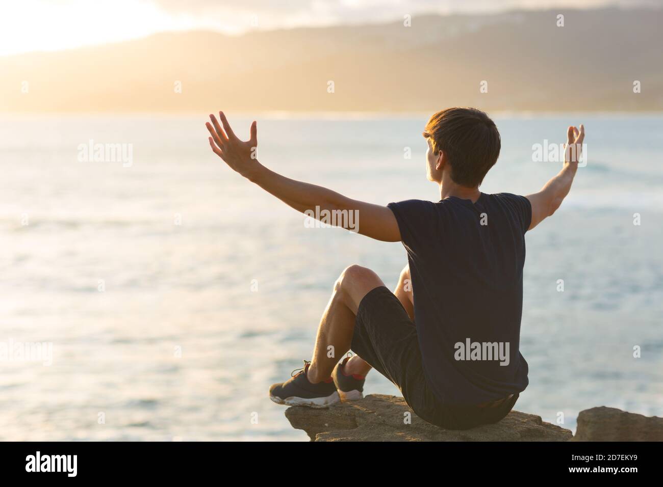 Happy young man feeling good sitting on top of a mountain cliff facing the ocean view and raising arms up to the sky. Hope and gratitude. Stock Photo
