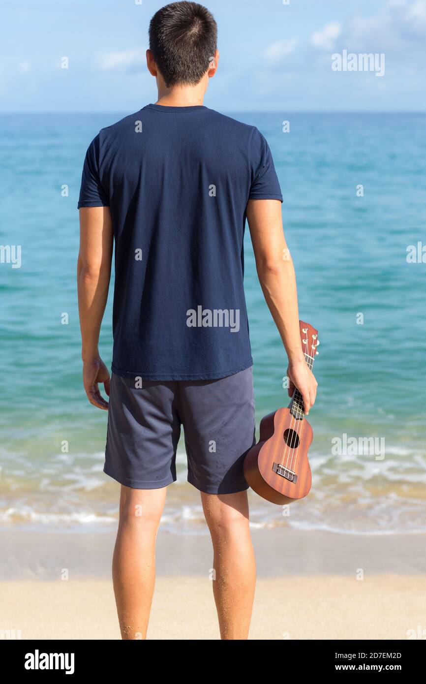 Young man holding an ukulele guitar standing on the beach facing the ocean and looking at the horizon. Stock Photo