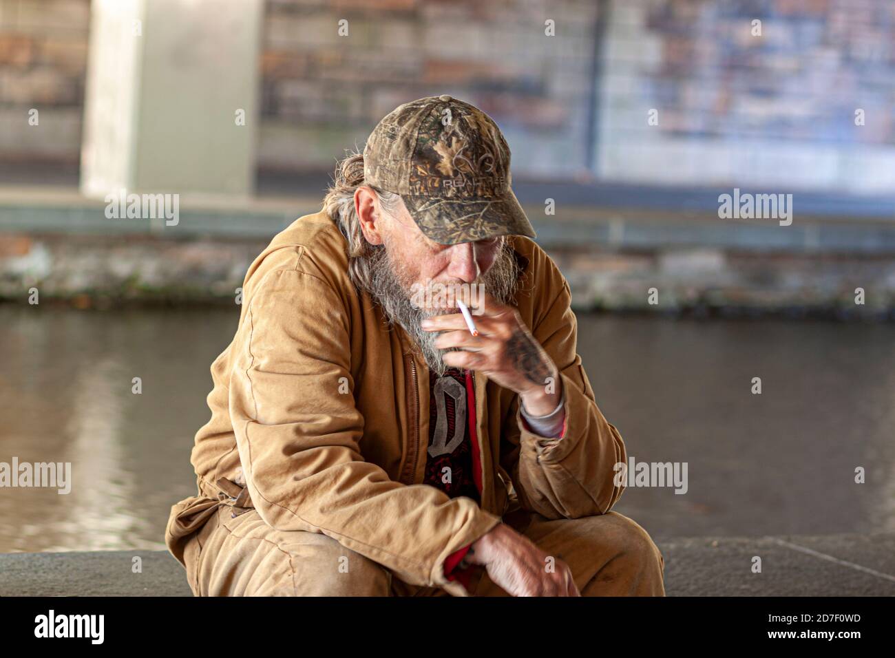 Frederick, MD, USA 10/14/2020: An elderly caucasian man with baseball hat and brown jacket is sitting by a river under bridge and smoking a cigarette. Stock Photo