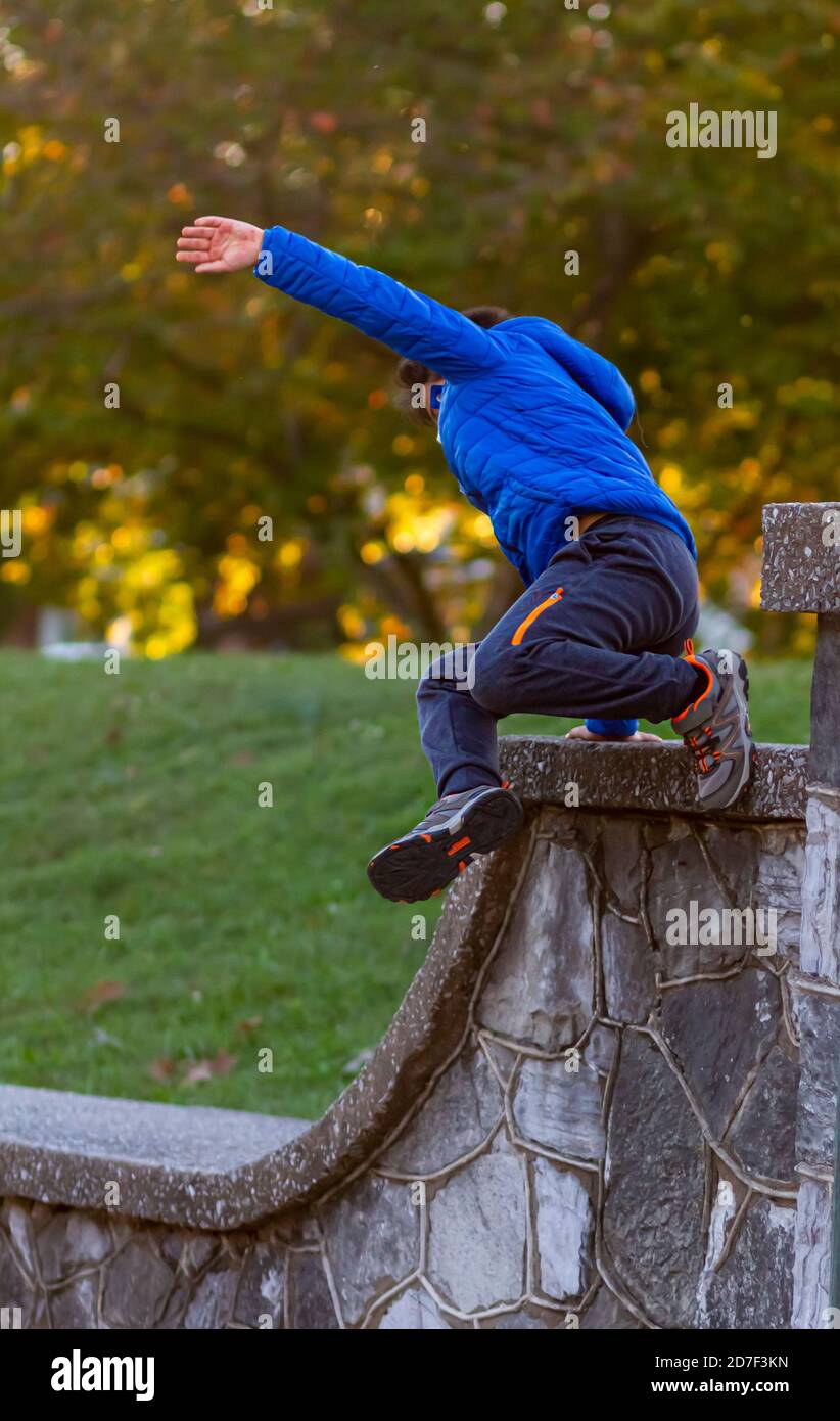 A hyperactive kid is jumping off a high wall as he is running away. The courageous boy wears track pants, sneakers and a hooded coat. He is full of en Stock Photo