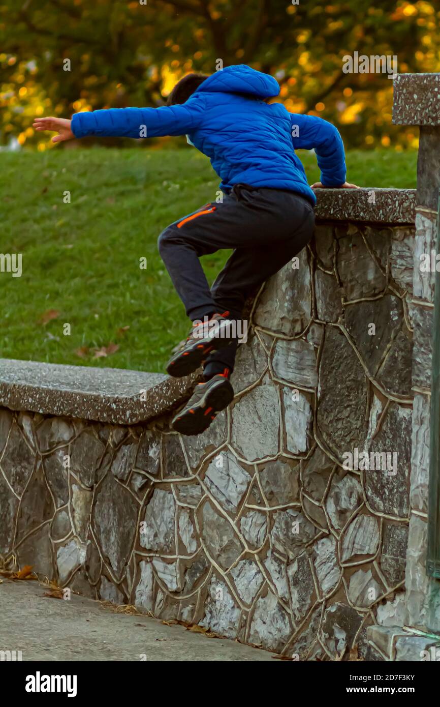 A hyperactive kid is jumping off a high wall as he is running away. The courageous boy wears track pants, sneakers and a hooded coat. He is full of en Stock Photo