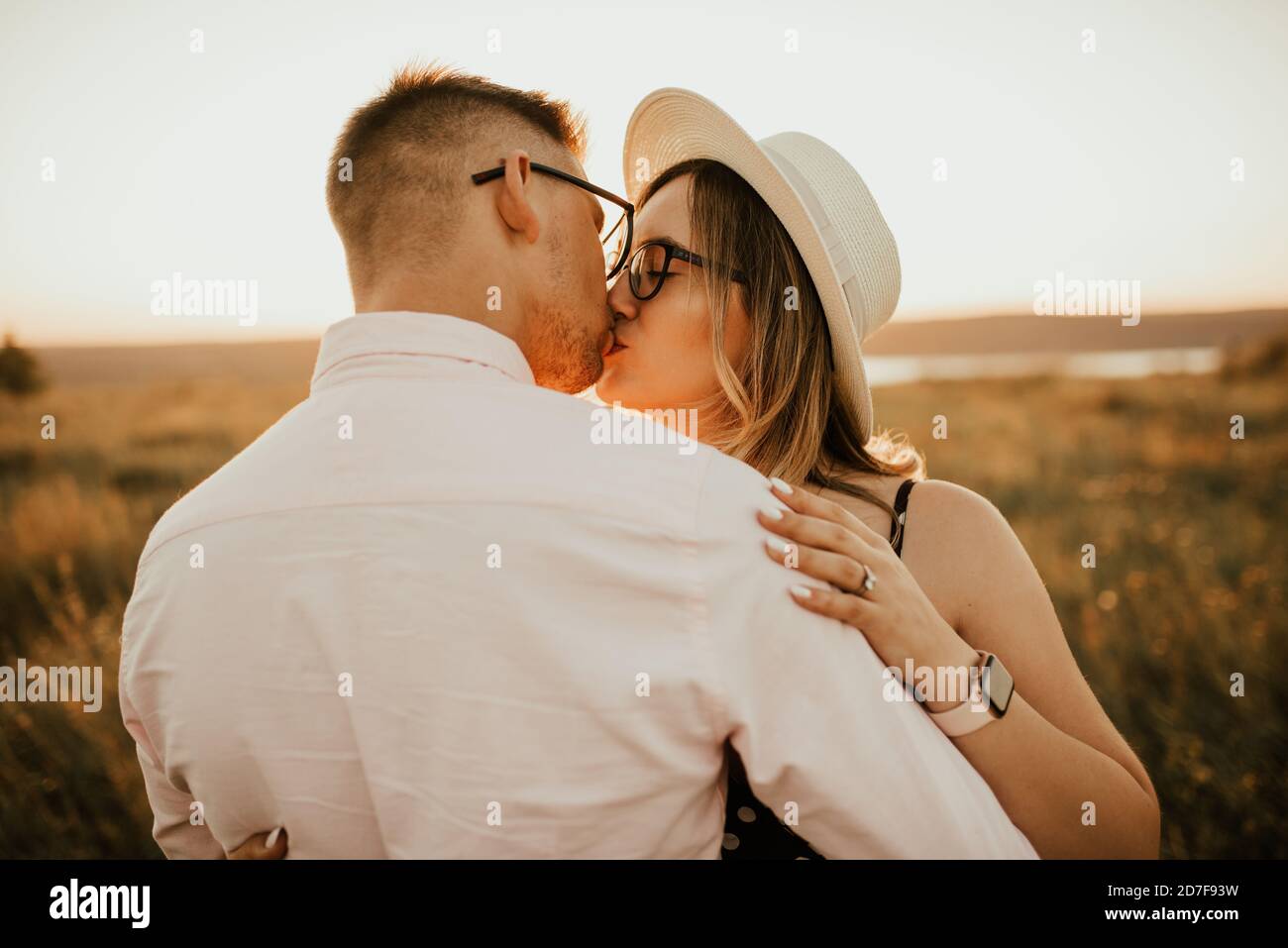 A man with a woman in a hat hug and kiss in the tall grass in the meadow Stock Photo
