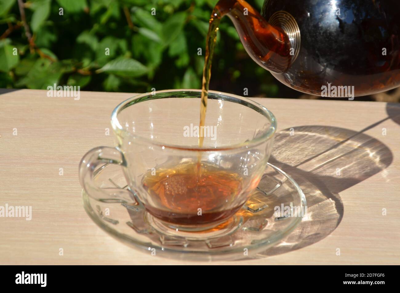 A girl pours tea from a transparent teapot into a transparent cup against a background of green foliage Good morning, outdoor cafe, energy boost. Tea Stock Photo
