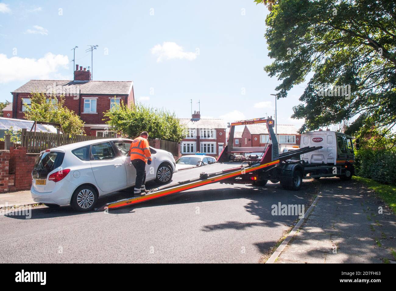 Man wearing high visability safety jacket loading car by winch onto hydraulic flatbed of recovery vehicle in restricted space Stock Photo