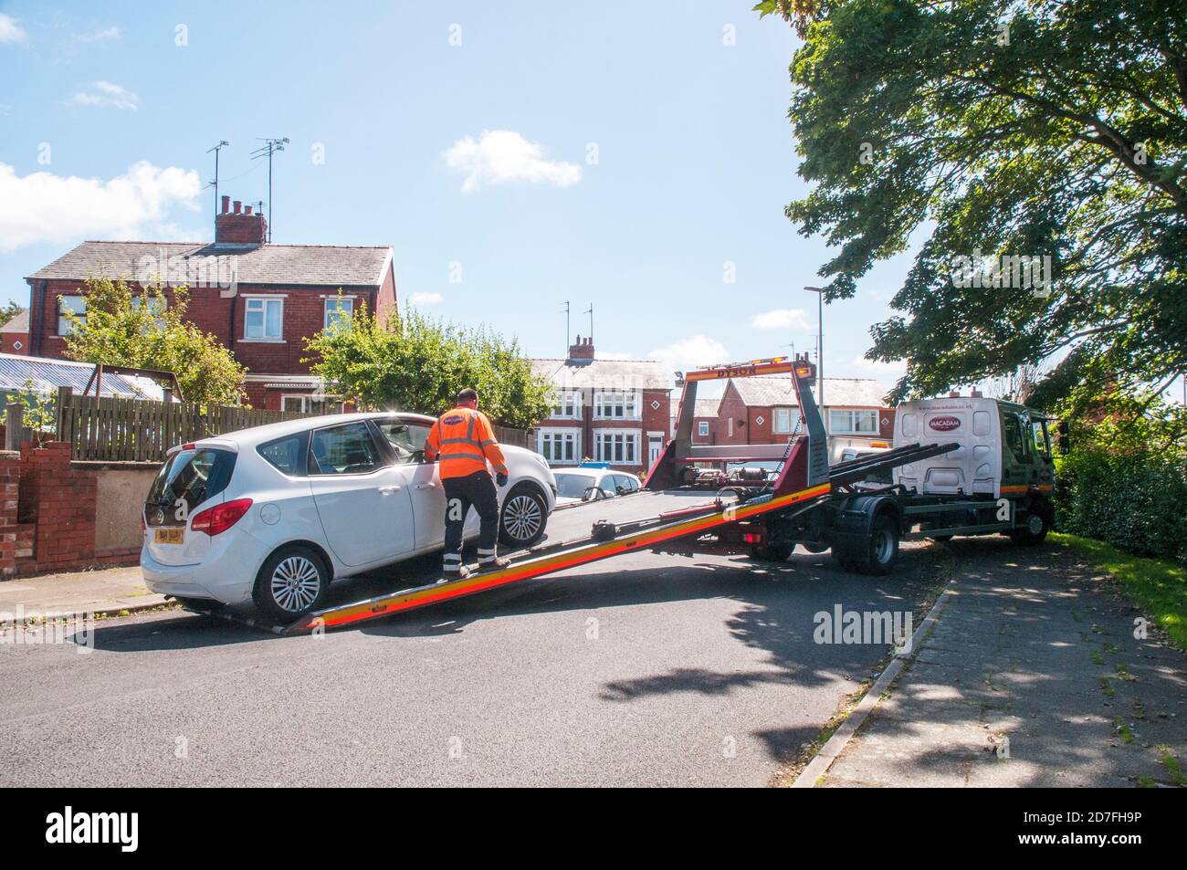 Man wearing high visability safety jacket loading car by winch onto hydraulic flatbed of recovery vehicle in restricted space Stock Photo