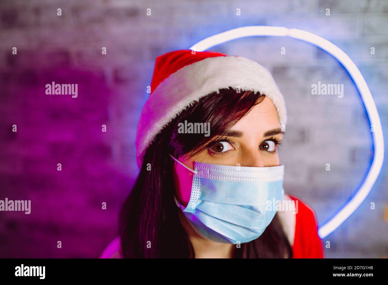Young woman in medical mask and Santa Claus hat against illuminated wall. Close up of brunette in Christmas hat and protective mask. Concept of safe Stock Photo