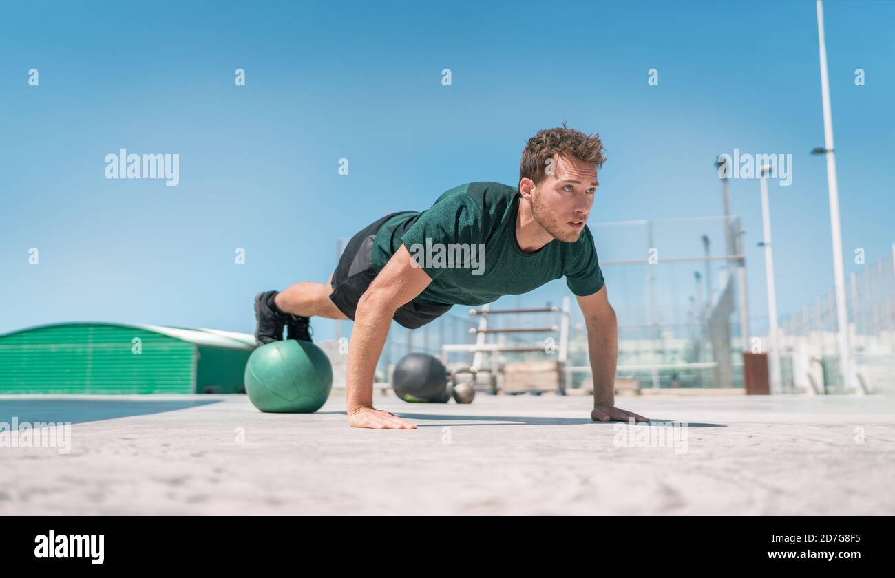 Fit exercises man strength training core doing balance push-ups workout at outdoor gym balancing on stability medicine ball with legs. Bodyweight Stock Photo