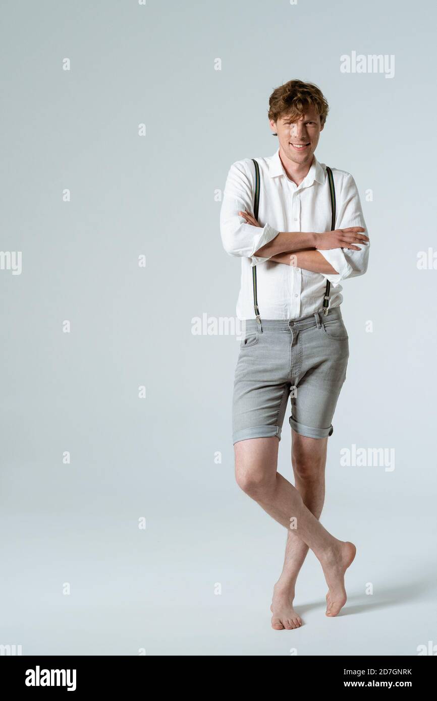 Stylish man in suspenders crossed his arms standing barefoot in full height in studio against white background. Charming dude Stock Photo