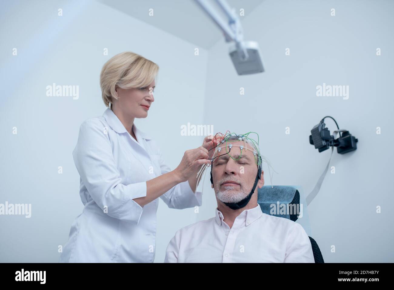 Female neurologist placing electrode cap on gray-haired male patient head Stock Photo