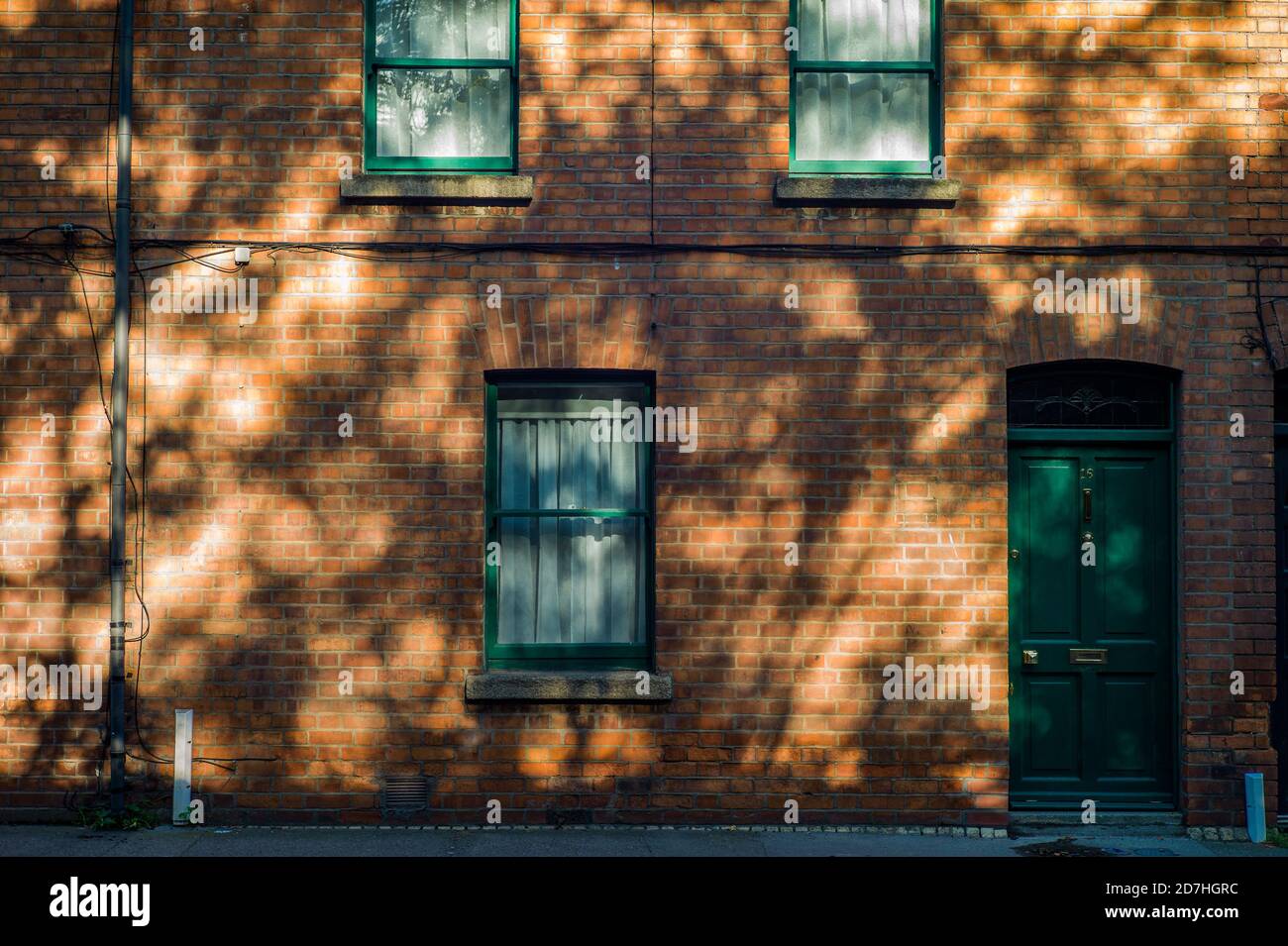 Facade of red brick house in South Dublin City. Dappled light on red brick from sunlight streaming through the branches of a tree. Stock Photo
