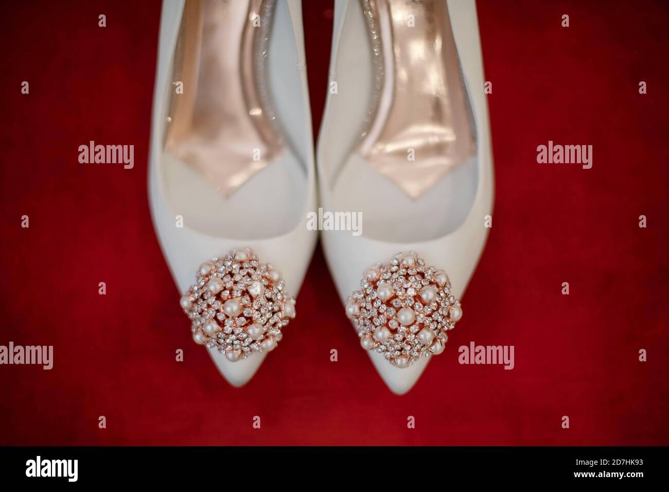 Close-up of beautiful white bridal pair of shoes embellished with pearls and crystal beads, set on a vivid red cushion, elegant leather footwear Stock Photo