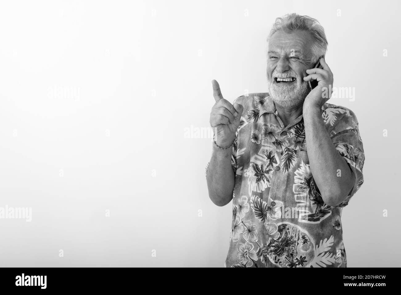 Studio shot of thoughtful happy senior bearded tourist man smiling and laughing while talking on mobile phone Stock Photo