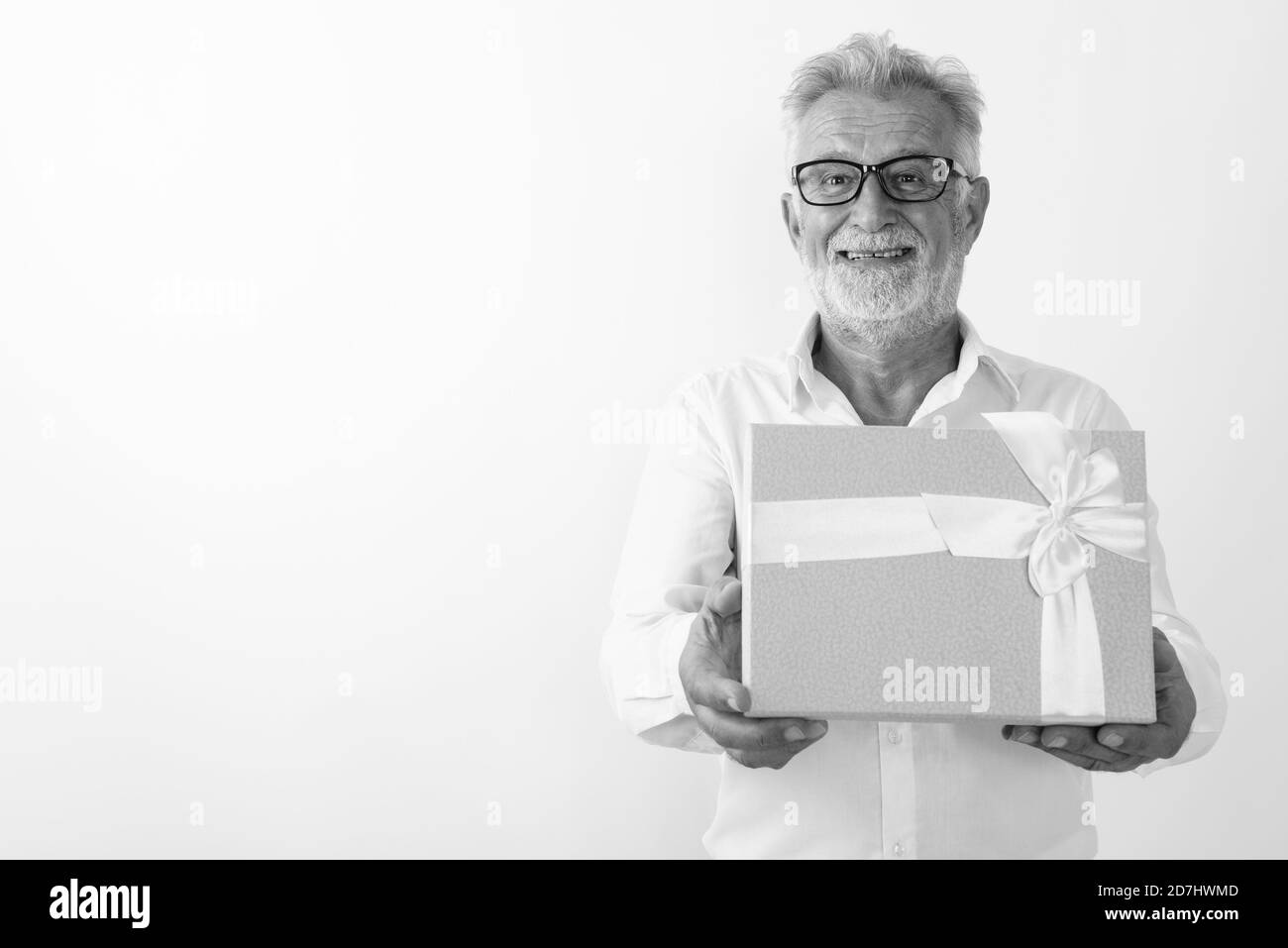 Studio shot of happy senior bearded man smiling and giving gift box while wearing eyeglasses against white background Stock Photo