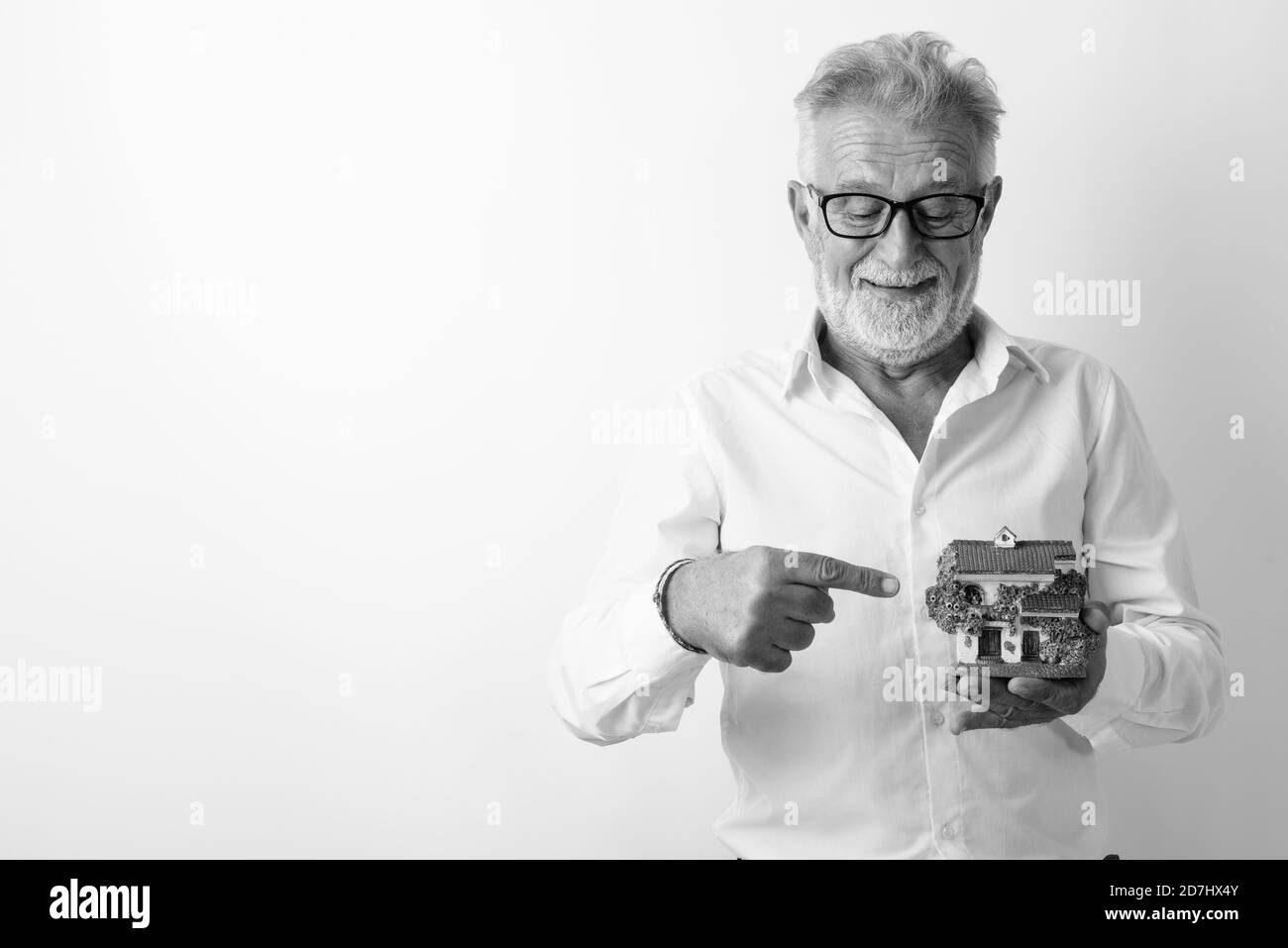 Studio shot of happy senior bearded man smiling while looking and pointing at house figurine with eyeglasses against white background Stock Photo