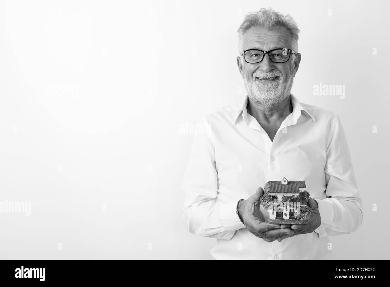 Studio shot of happy senior bearded man smiling while holding house figurine and wearing eyeglasses against white background Stock Photo