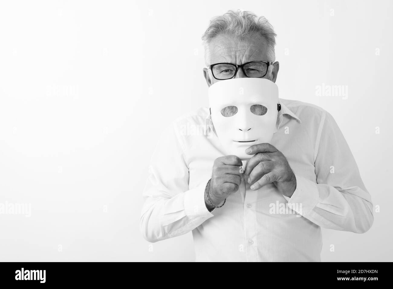 Studio shot of handsome senior bearded man covering mouth with white mask while wearing eyeglasses against white background Stock Photo