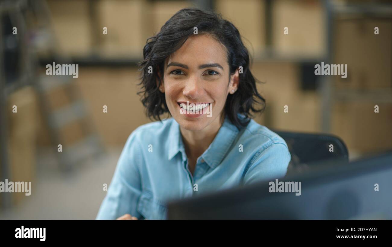 Female Inventory Manager Works Sitting at Her Desk and Smiling at the Camera. In the Background Warehouse Storeroom with Shelves Full of Cardboard Box Stock Photo