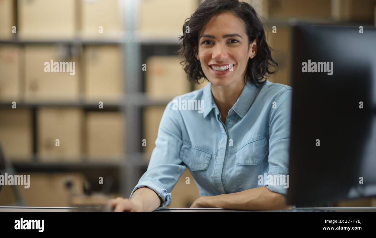 Female Inventory Manager Works Sitting at Her Desk, Working on Computer and Smiling at the Camera. In the Background Warehouse Storeroom with Shelves Stock Photo