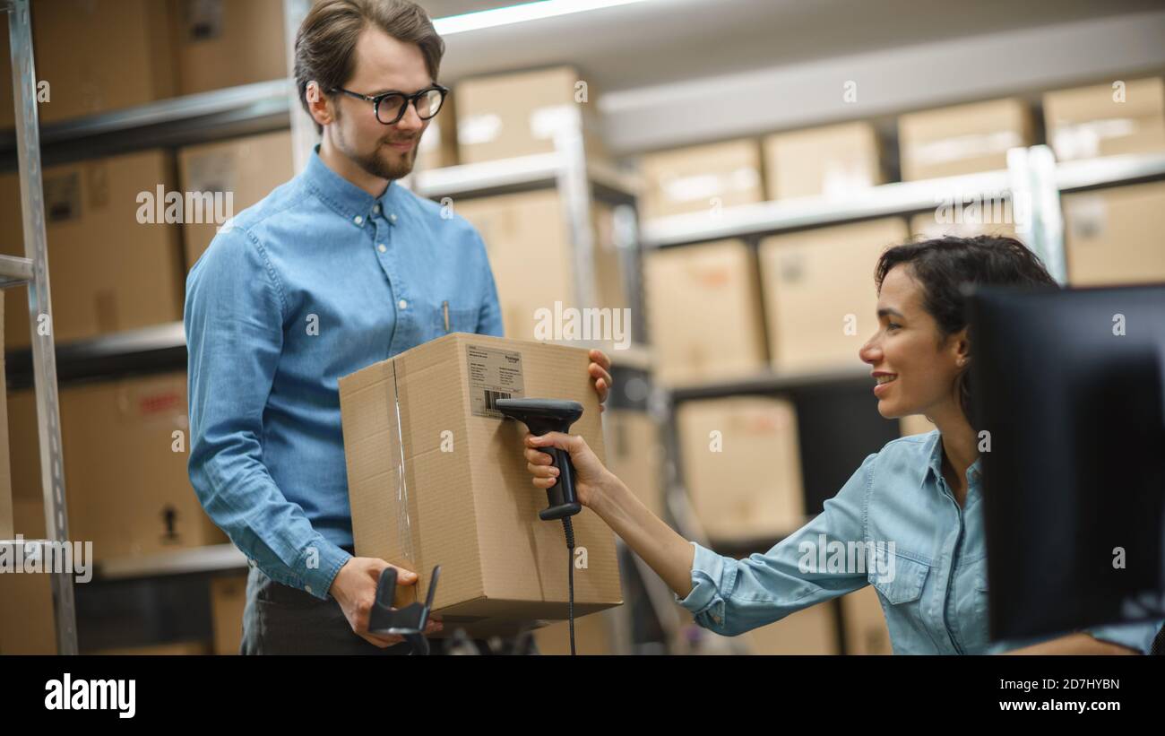 Female Inventory Manager Scans Cardboard Box and Digital Tablet with Barcode Scanner, Male Worker Holds Package. In the Background Rows of Cardboard Stock Photo