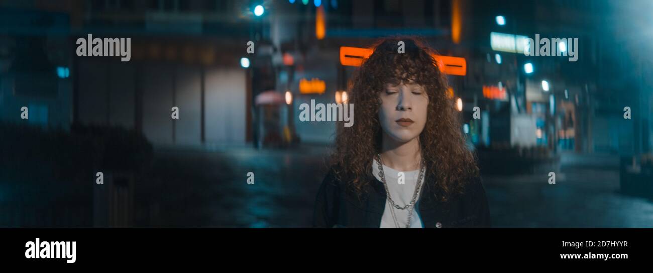 Curly hair woman in a white t-shirt and black jacket leaves a wonderful street with red led lights at the night and she is turns and starts walking, f Stock Photo