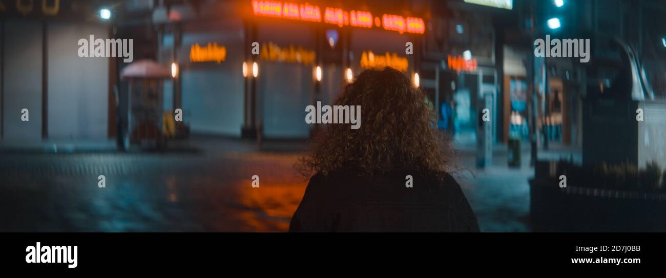 Curly hair woman in a white t-shirt and black jacket leaves a wonderful street with red led lights at the night and she is turns and starts walking, f Stock Photo