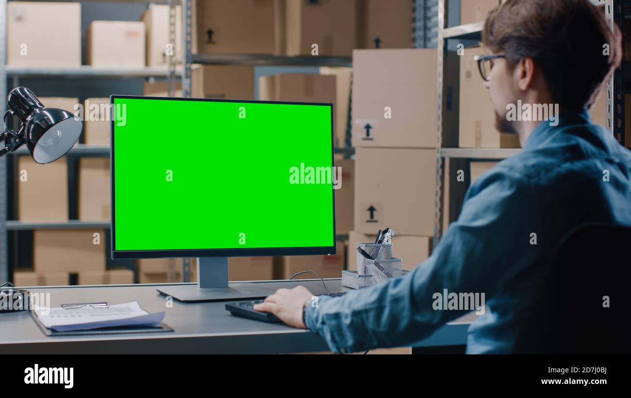 Warehouse Inventory Manager Works on a Green Mock-up Screen Computer while Sitting at His Desk. In the Background Shelves Full of Cardboard Box Stock Photo