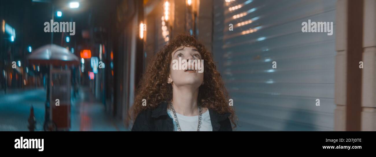 Stressful unhappy young attractive woman with curly hair in white t-shirt and black jacket is walking on the street at the night, fashion clothing and Stock Photo