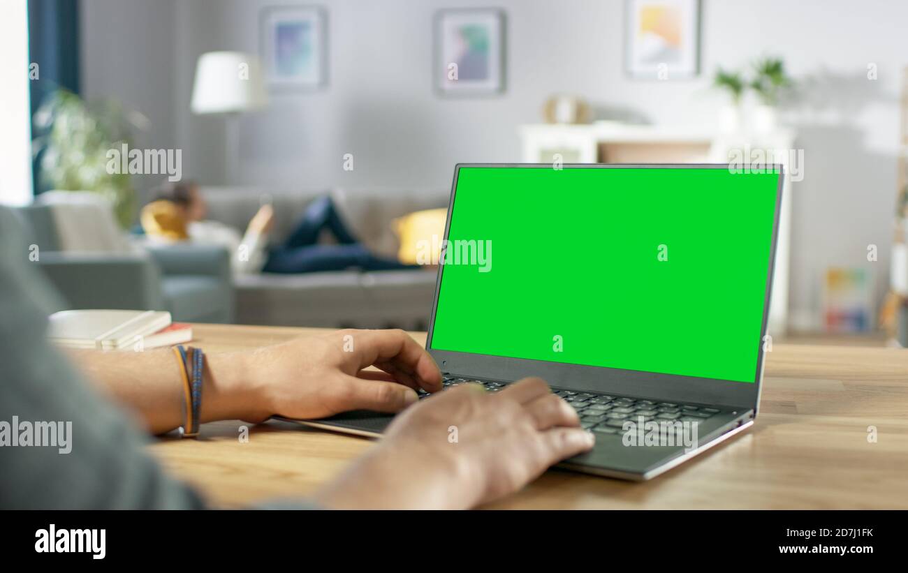 Over the Shoulder Shot of Mans Hands Typing on a Laptop with Green Mock-up Screen. In the Background Cozy Living Room with Woman Relaxing on a Sofa. Stock Photo
