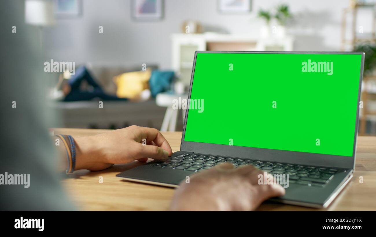 Over the Shoulder Shot of Mans Hands Using Laptop with Green Mock-up Screen. In the Background Cozy Living Room with Woman Relaxing on a Sofa. Stock Photo