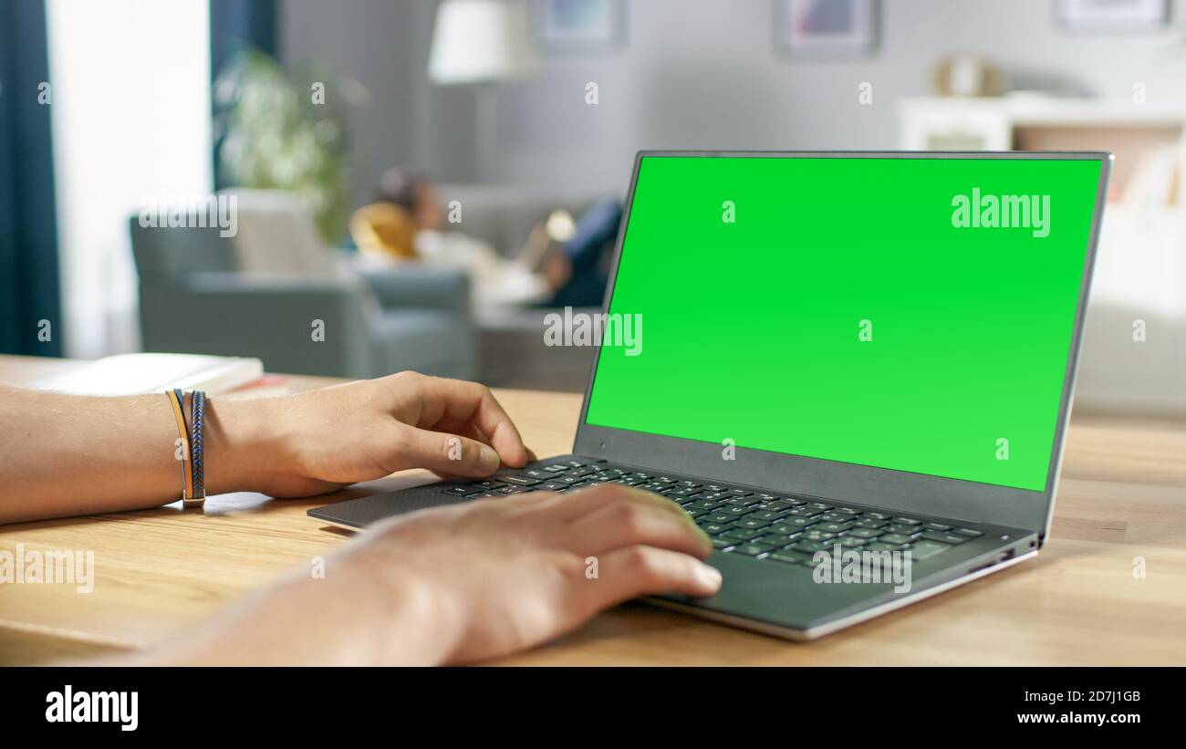 Over the Shoulder Shot of Mans Hands Typing on a Laptop with Green Mock-up Screen. In the Background Cozy Living Room with Woman Relaxing on a Sofa. Stock Photo