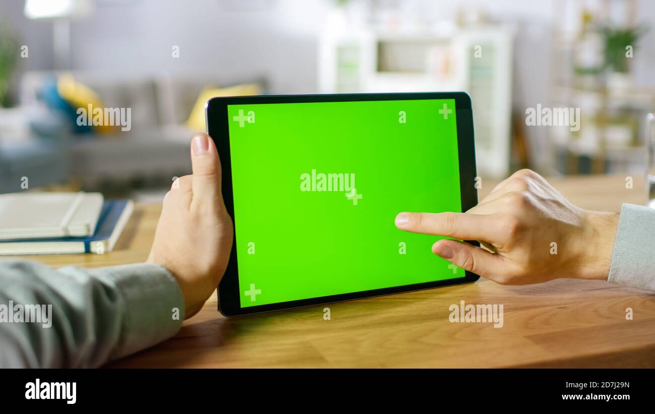 Close-up of Man Using Hand Gestures on Green Mock-up Screen Digital Tablet Computer in Landscape Mode while Sitting at His Desk. In the Background Stock Photo