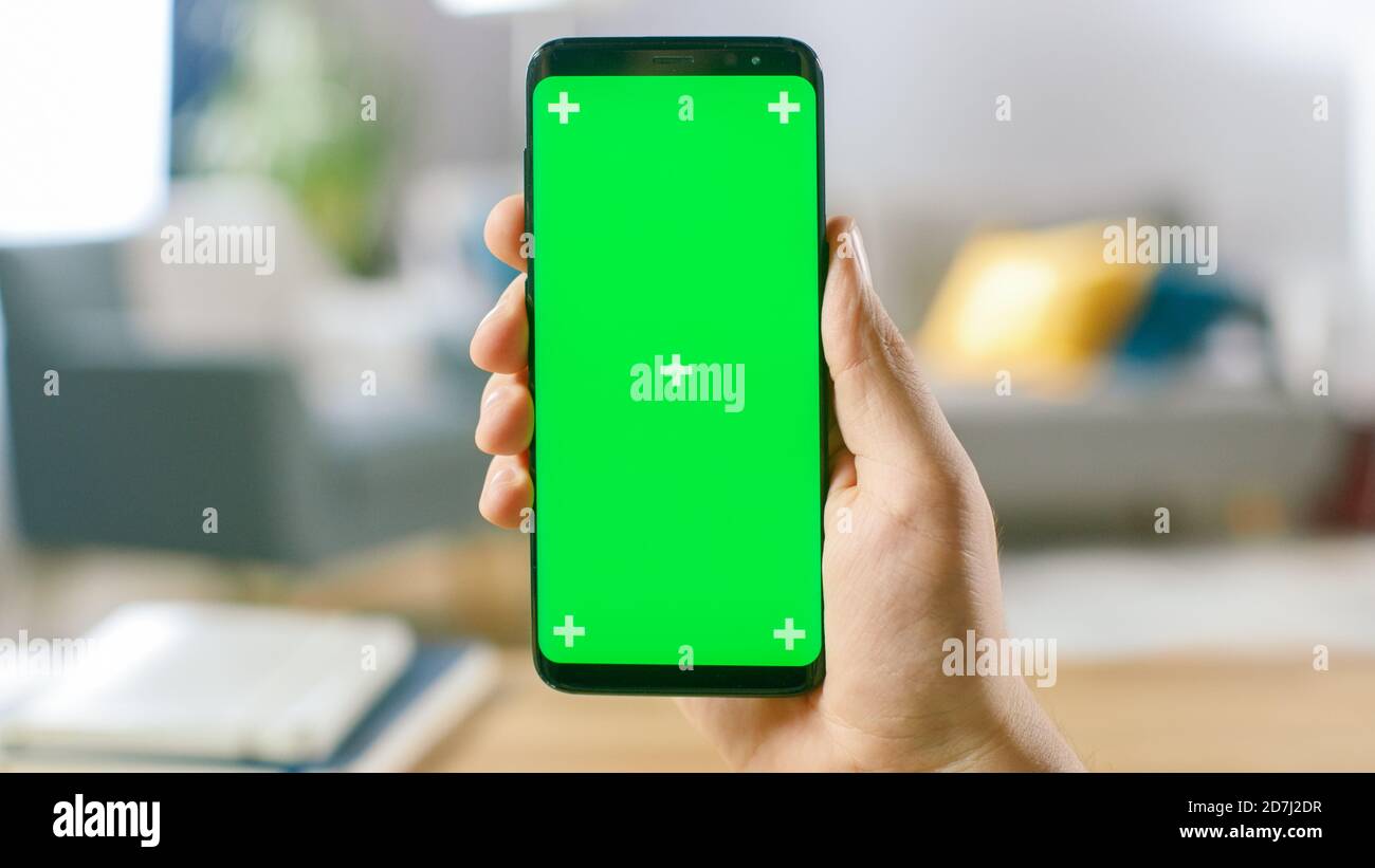Close-up of a Man's Hand Holding Green Mock-up Screen Smartphone. Modern Mobile Phone. In the Background Cozy Living Room or Home Office. Stock Photo
