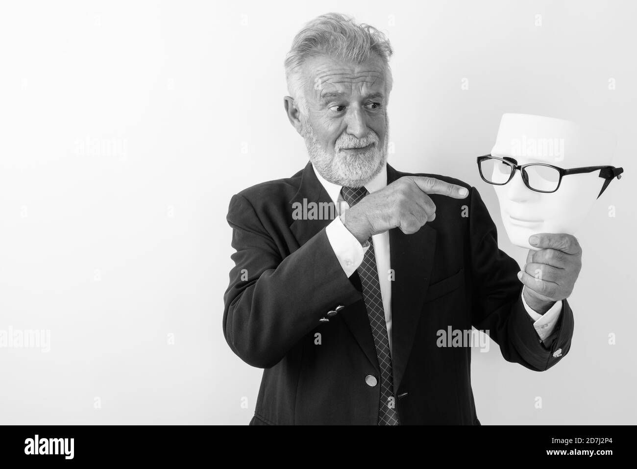 Studio shot of handsome senior bearded businessman looking and pointing at white mask with eyeglasses against white background Stock Photo