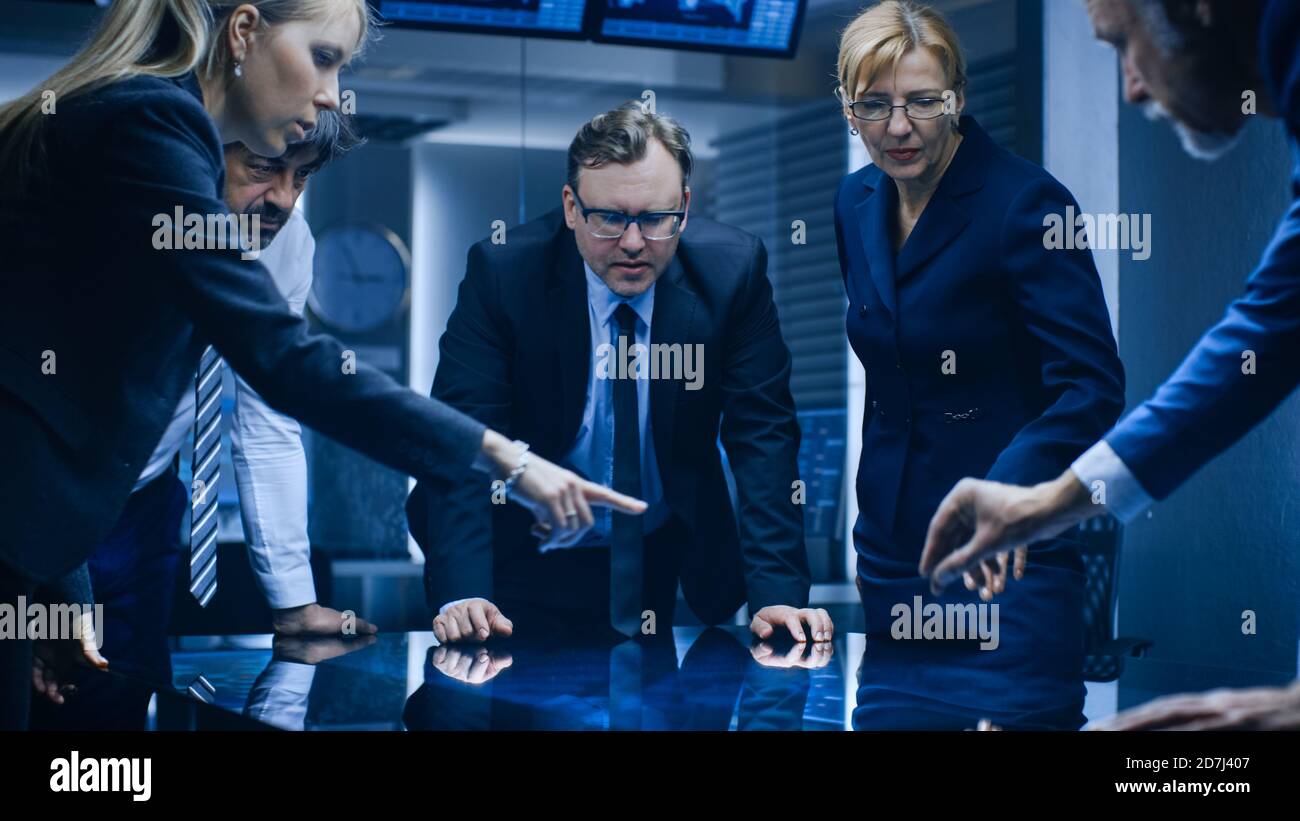 Diverse Team of Government Intelligence Agents Standing Around Digital Touch Screen Table and Tracking Suspect. Big Dark Surveillance Room Full of Stock Photo