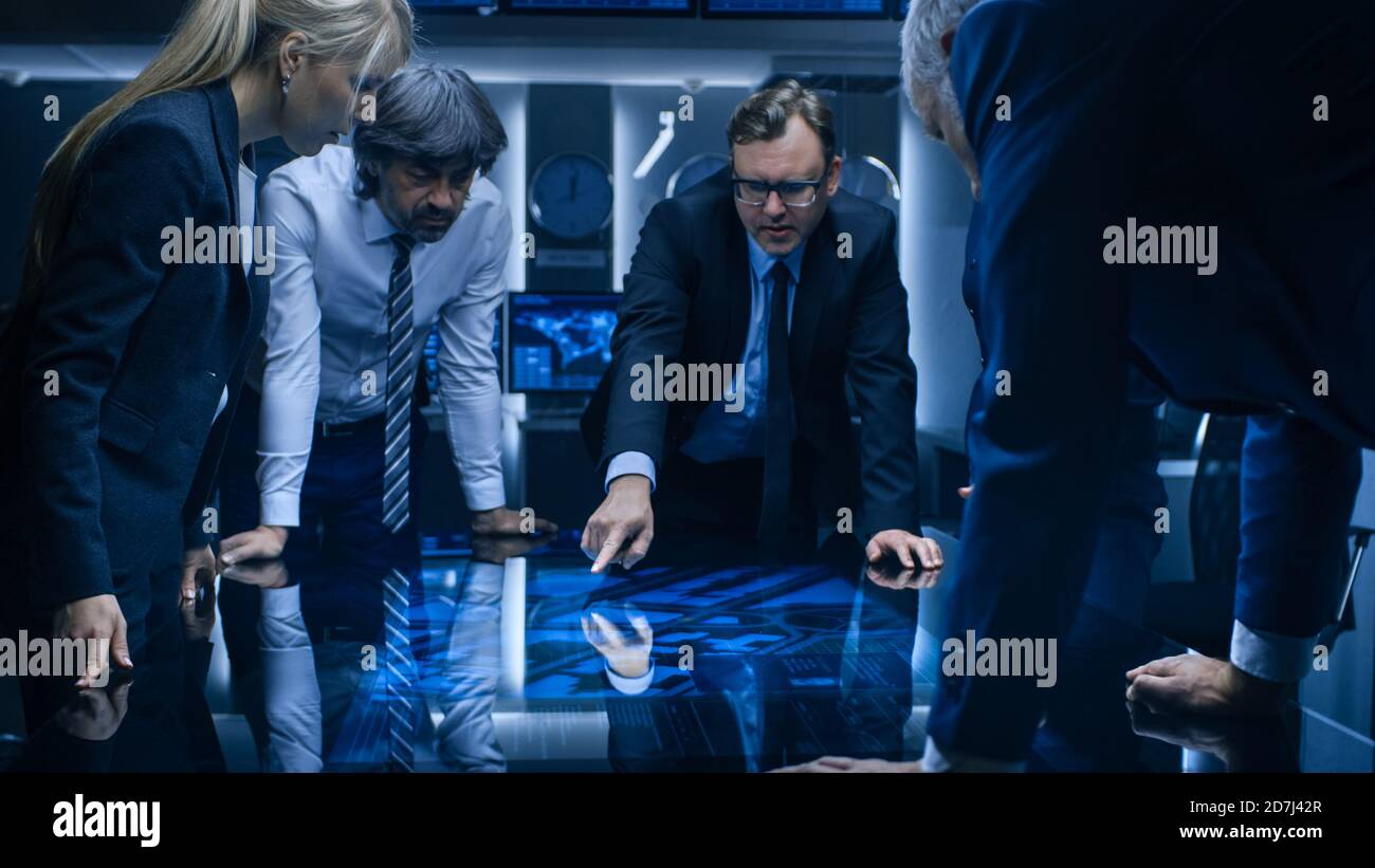 Diverse Team of Government Intelligence Agents Standing Around Digital Touch Screen Table and Tracking Suspect. Big Dark Surveillance Room Full of Stock Photo