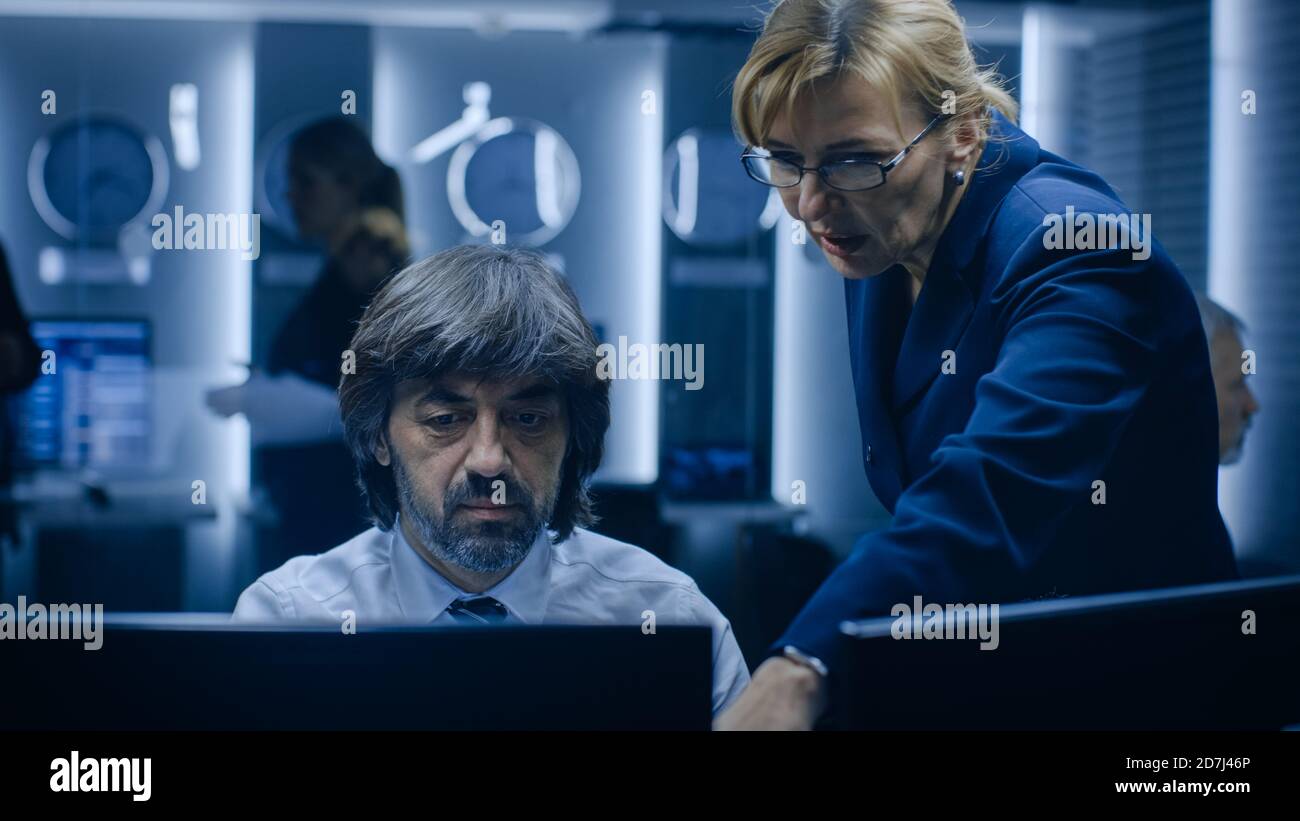 Female Manager Talks with Professional Operator Working on a Personal Computer in the System Monitoring Room. High Profile Specialist Working. Stock Photo