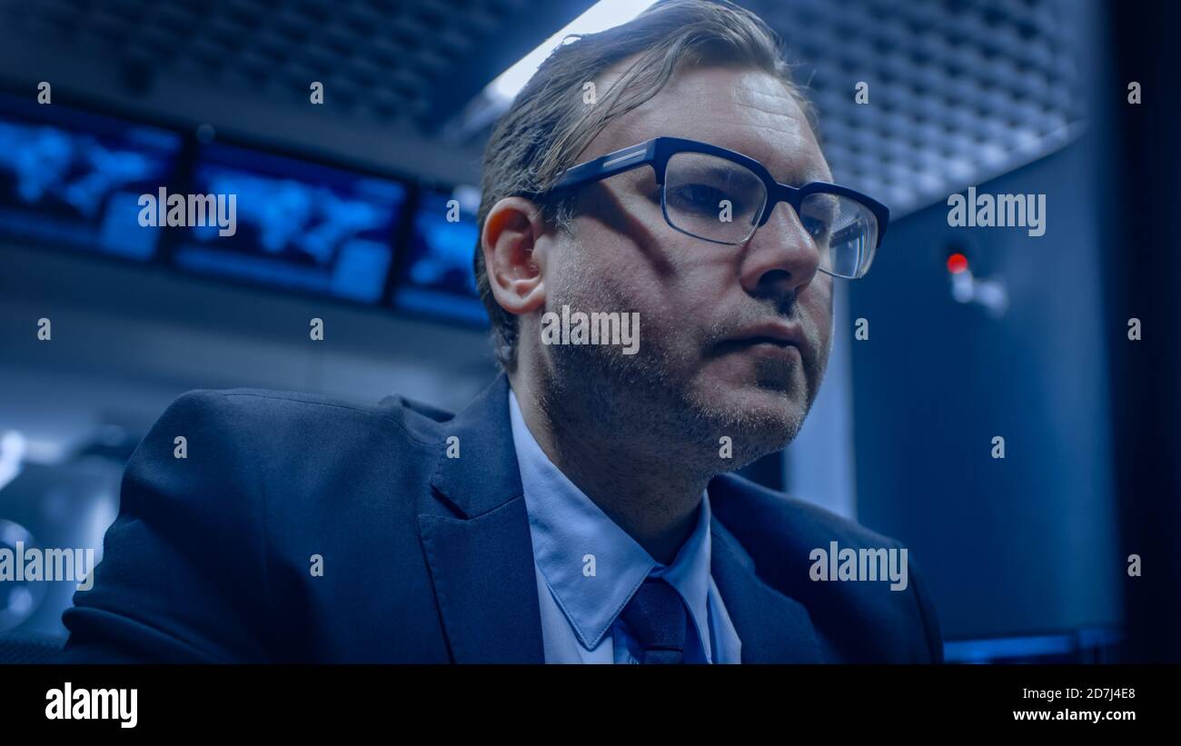 Portrait of the Serious Specialist Working on Computer in the Monitoring Control Room with Teams Solving Problems in the Background. Stock Photo
