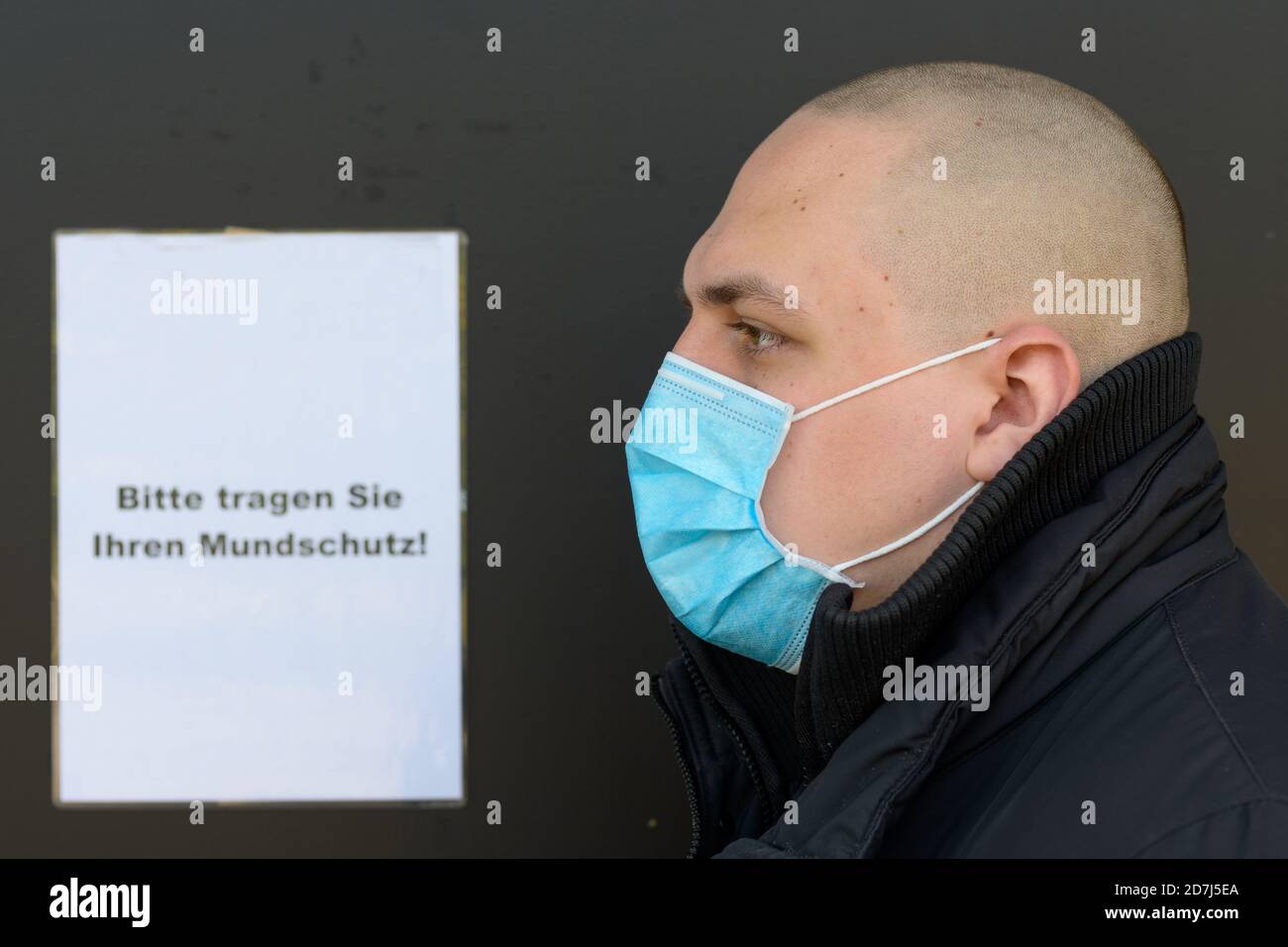 Profile of a young man wearing a face mask for infection control during the Covid-19 pandemic standing in front of a German text sign instructing peop Stock Photo