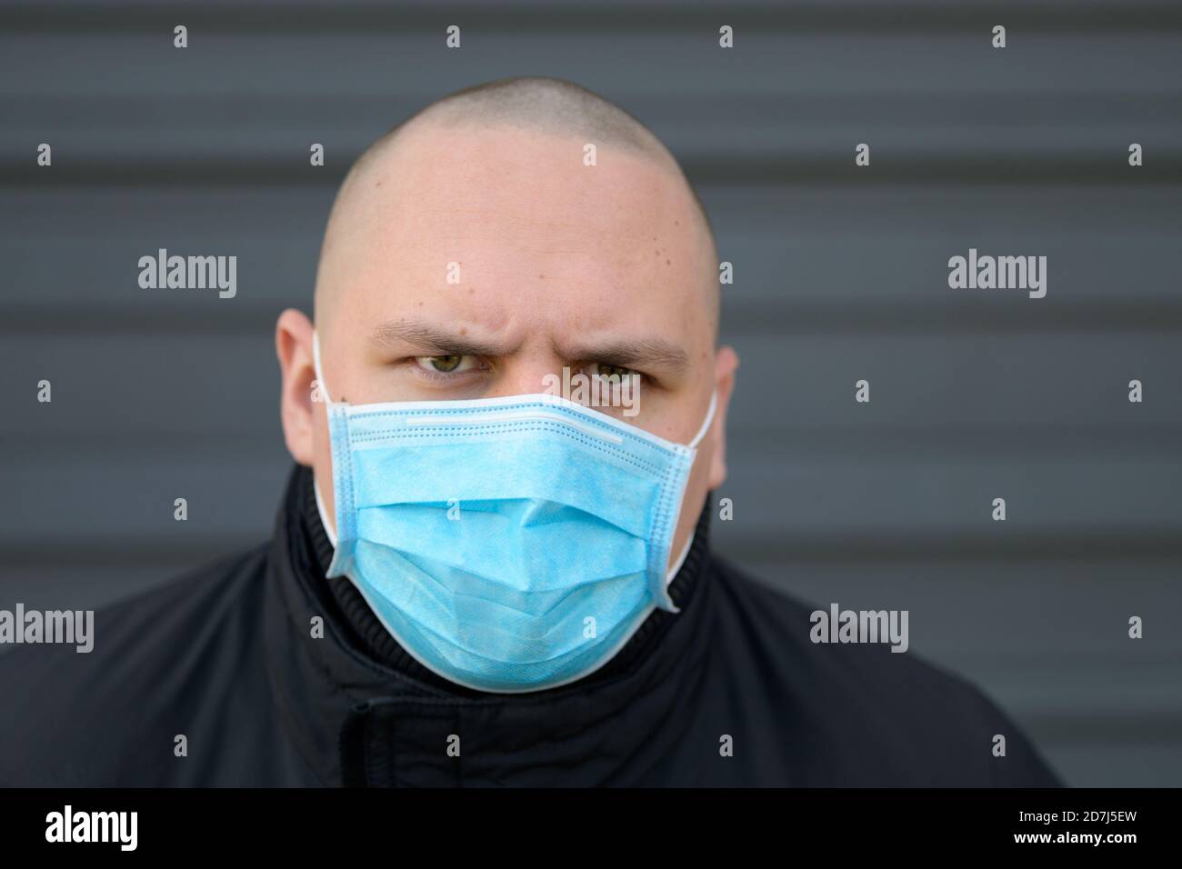 Angry young man wearing a surgical face mask frowning as he looks with a determined expression during the Covid-19 pandemic against a grey exterior wa Stock Photo