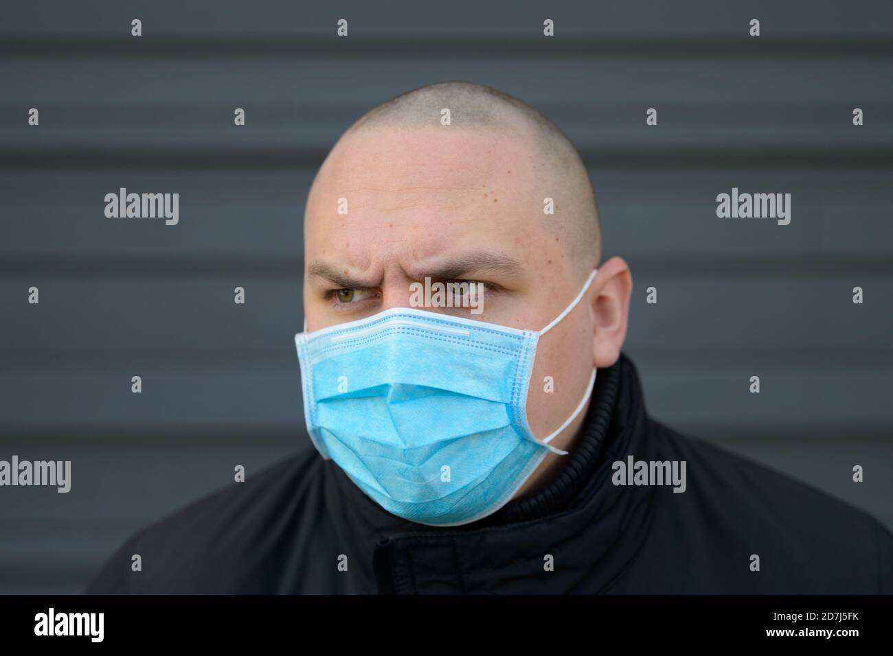 Angry young man wearing a surgical face mask frowning as he looks away with a determined expression during the Covid-19 pandemic against a grey exteri Stock Photo