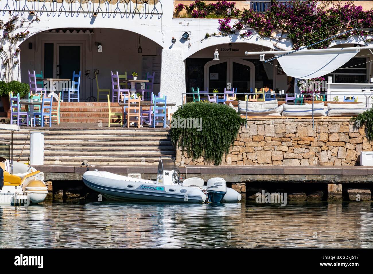 Marina dell' Orso di Poltu Quatu Waterfront. Quayside View of Hotel and Restaurant with Colourful Chairs and Moored Dinghy. Sardinia, Italy. Stock Photo