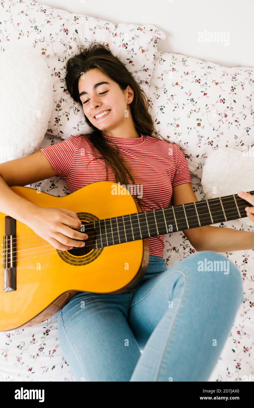 Young woman lying on back while playing guitar in bedroom Stock Photo