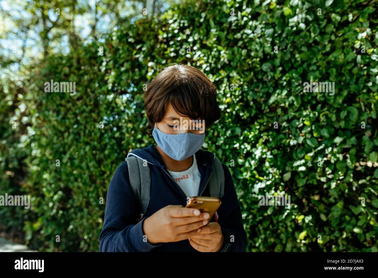 Schoolboy wearing mask using mobile phone while standing by plants Stock Photo