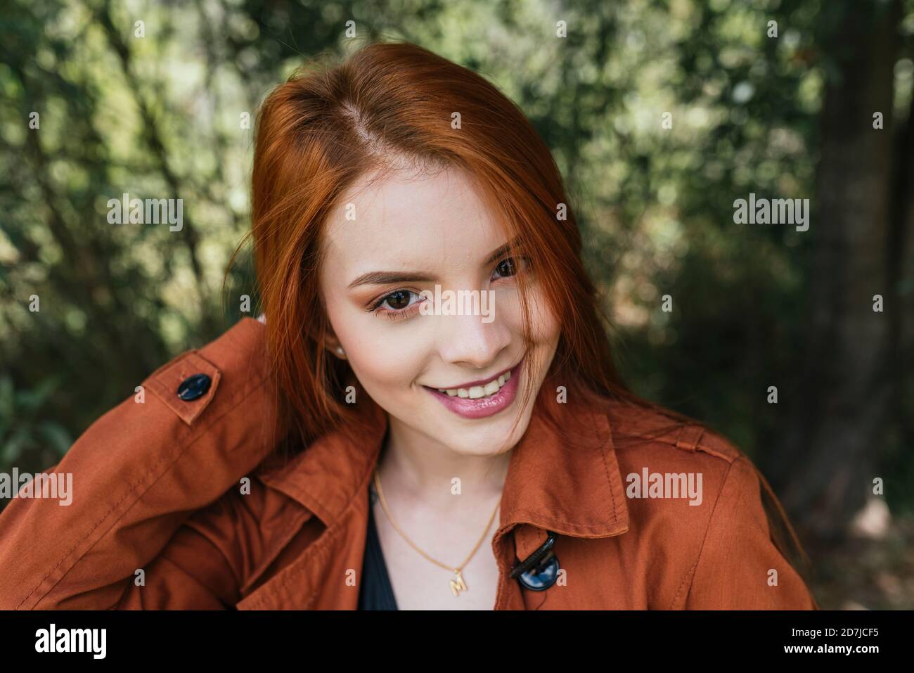 Smiling beautiful young redhead woman wearing brown jacket at park Stock Photo