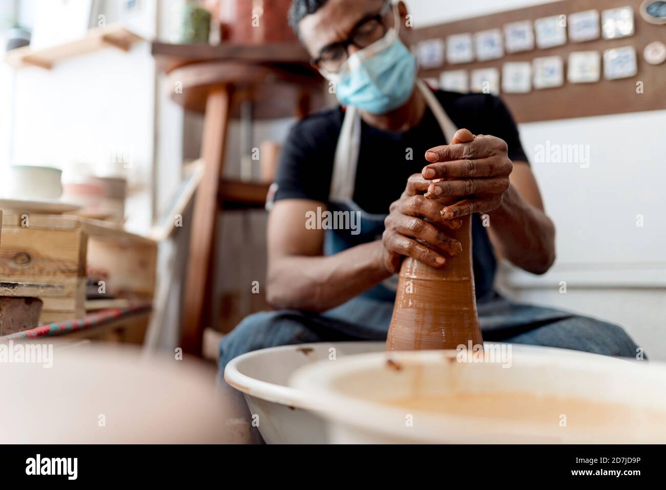 Male potter wearing mask molding shape to clay while sitting in workshop Stock Photo